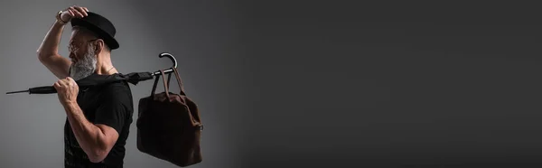 Side view of stylish senior man in derby hat holding umbrella with leather bag on grey, banner — Stock Photo