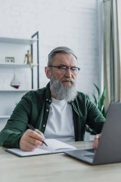 Bärtiger älterer Mann mit Stift und Laptop bei der Arbeit von zu Hause aus — Stockfoto