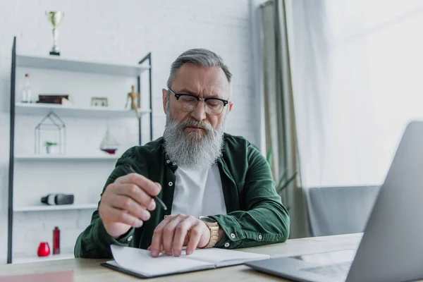 Бородатый мужчина держит ручку и смотрит на ноутбук рядом с ноутбуком на столе — стоковое фото