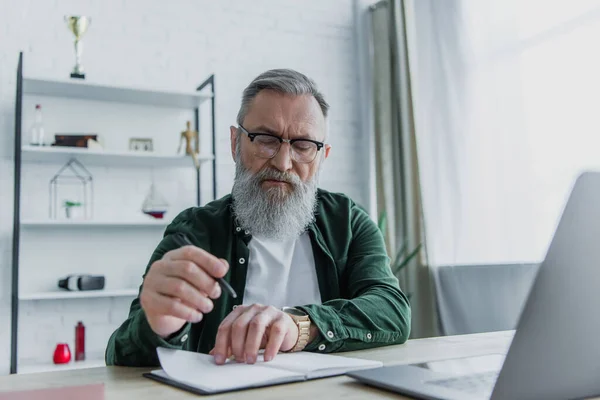 Barbudo hombre mayor sosteniendo pluma y mirando portátil cerca de la computadora portátil en el escritorio - foto de stock