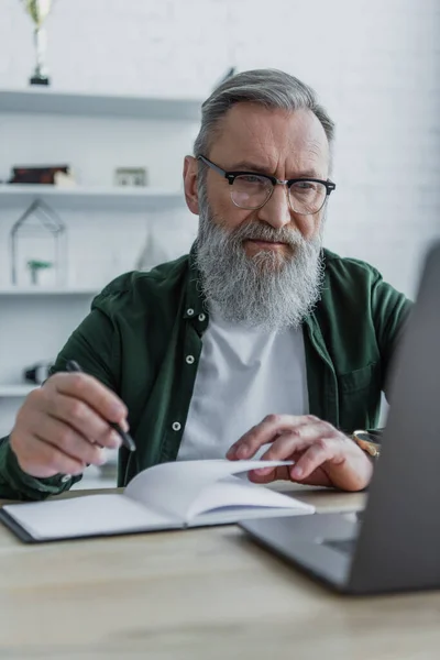 Homme âgé barbu tenant stylo près d'un ordinateur portable et en utilisant un ordinateur portable tout en travaillant à la maison — Photo de stock