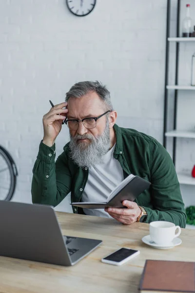Uomo anziano pensoso e barbuto in occhiali da vista che tiene la penna e utilizza il computer portatile mentre lavora da casa — Foto stock