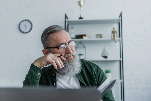 Пеніс і бородатий старший чоловік в окулярах тримає ручку і дивиться геть, працюючи з дому — стокове фото