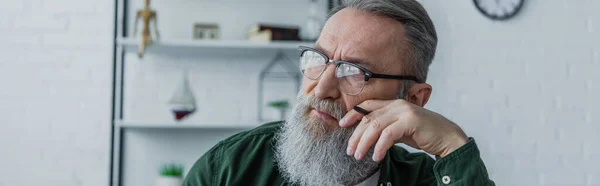 Задумчивый и бородатый мужчина в очках держит ручку и смотрит в сторону дома, баннер — стоковое фото
