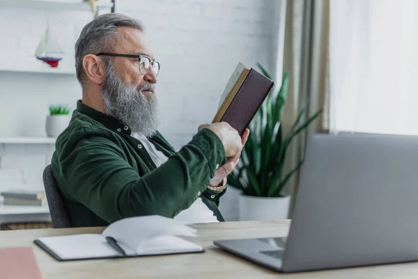 Barbuto uomo anziano in occhiali da vista lettura libro vicino laptop sulla scrivania — Foto stock