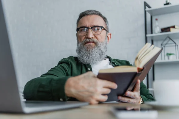 Uomo anziano barbuto in occhiali guardando la fotocamera vicino al computer portatile mentre tenendo libro — Foto stock