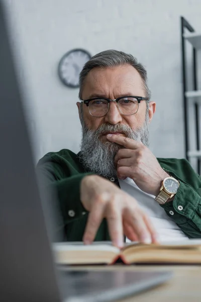 Nachdenklicher bärtiger Senior mit Brille blickt auf Laptop, während er Buch mit der Hand berührt — Stockfoto