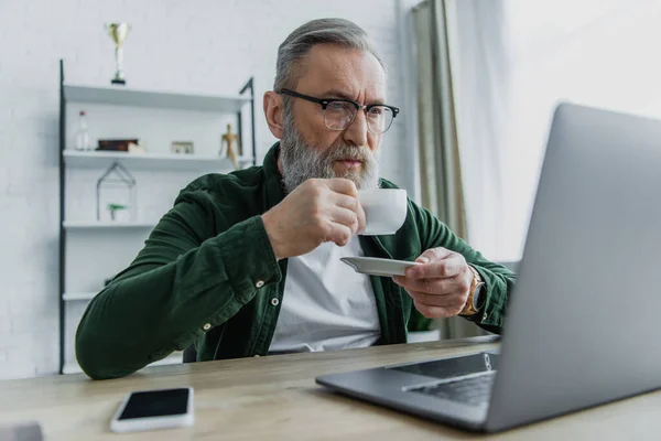Bärtiger älterer Mann mit Brille trinkt Kaffee und schaut von zu Hause aus auf den Laptop — Stockfoto