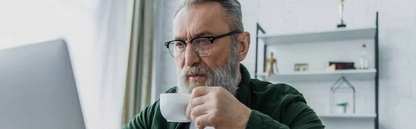 Uomo anziano barbuto in occhiali da vista bere caffè e guardando il computer portatile mentre si lavora da casa, banner — Foto stock