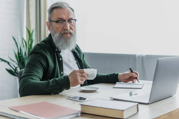 Бородатый мужчина в очках держит чашку кофе и ручку возле ноутбука во время работы из дома — стоковое фото