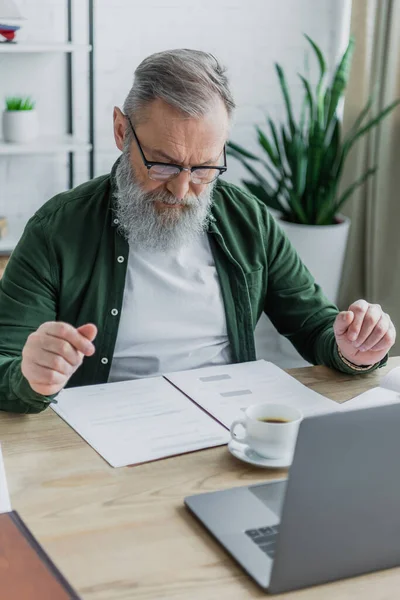 Bärtiger älterer Herr mit Brille schaut sich Dokumente in der Nähe von Kaffee und Laptop an, während er von zu Hause aus arbeitet — Stockfoto