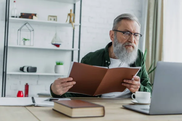 Bärtiger älterer Mann mit Brille hält Ordner in der Hand und blickt auf Laptop auf Schreibtisch — Stockfoto