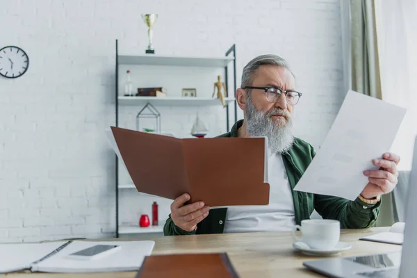 Senior mit Brille hält Aktenmappe in der Hand und schaut Dokument neben Laptop auf Schreibtisch — Stockfoto
