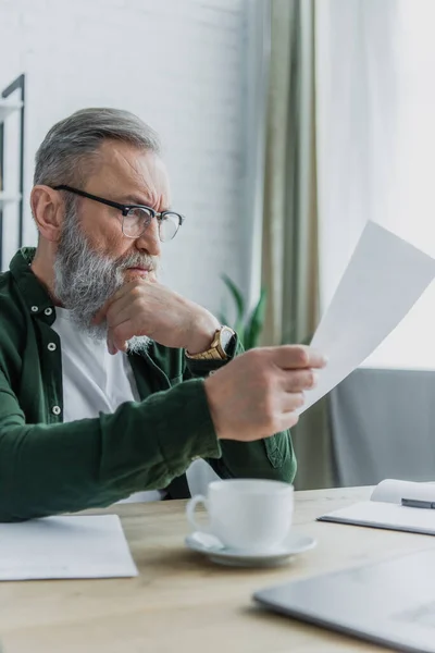 Uomo anziano in occhiali da vista guardando documento vicino tazza e laptop offuscata sulla scrivania — Foto stock