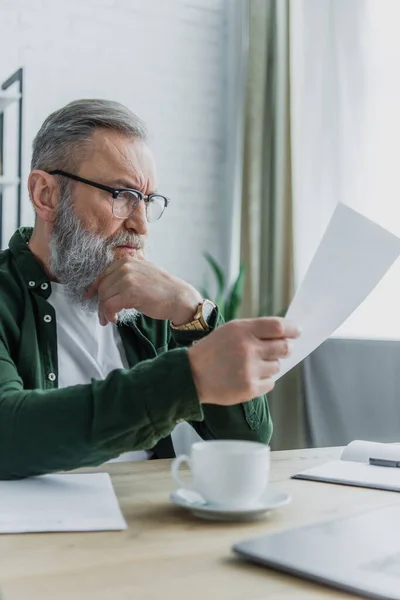 Senior en lunettes regardant le document près de tasse et ordinateur portable flou sur le bureau — Photo de stock