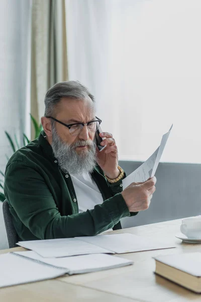 Bearded senior man in eyeglasses talking on smartphone and holding document — Stock Photo