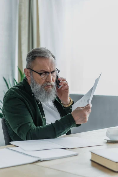 Bärtiger Senior mit Brille spricht auf Smartphone und hält Dokument in der Hand — Stockfoto