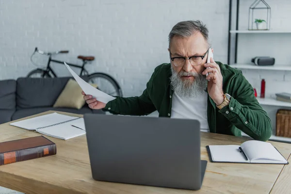 Homme âgé mécontent dans les lunettes parler sur smartphone et tenant le document près de l'ordinateur portable sur le bureau — Photo de stock