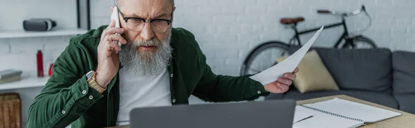 Незадоволений старший чоловік в окулярах розмовляє на смартфоні і тримає документ біля ноутбука, банер — стокове фото