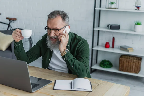 Hombre mayor barbudo en gafas con taza y hablando en el teléfono inteligente cerca de la computadora portátil - foto de stock