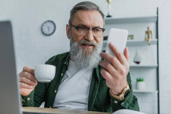 Бородатый мужчина в очках держит чашку и смотрит на смартфон — стоковое фото