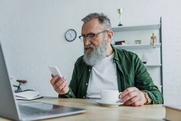 Bärtiger älterer Mann mit Brille hält Tasse in der Hand und blickt auf sein Smartphone neben dem Laptop auf dem Schreibtisch — Stockfoto