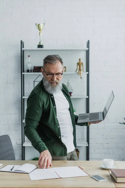 Homme âgé barbu dans des lunettes debout avec ordinateur portable tout en regardant les papiers sur le bureau — Photo de stock