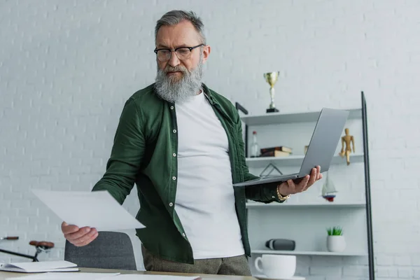 Bearded senior man in eyeglasses standing with laptop while looking at papers at home — Stock Photo