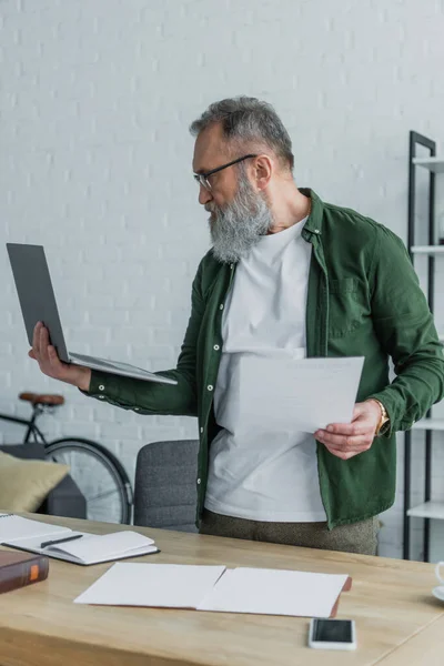 Uomo anziano barbuto in occhiali in piedi con computer portatile mentre in possesso di documento a casa — Foto stock