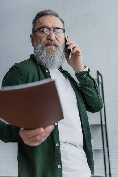 Hombre mayor barbudo en gafas graduadas de pie con carpeta mientras habla en el teléfono inteligente - foto de stock