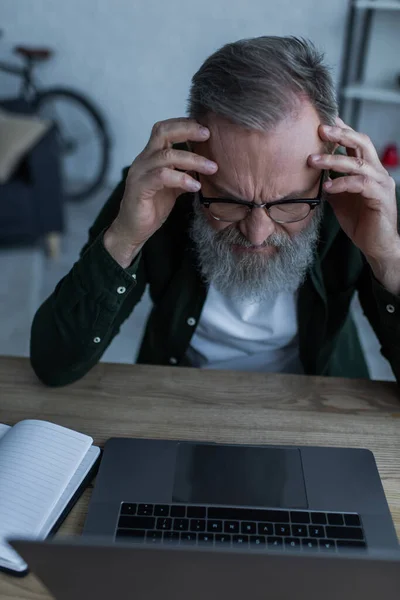 Estresado hombre mayor en gafas frunciendo el ceño cerca de la computadora portátil en casa - foto de stock