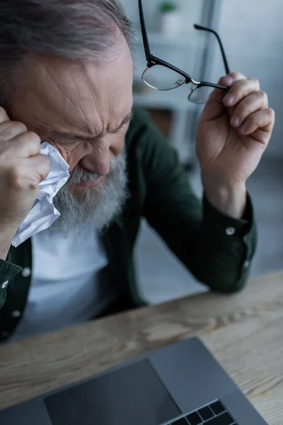 High angle view of bearded senior man holding eyeglasses and crumpled paper near laptop — Stock Photo