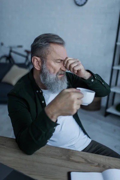 Cansado barbudo y hombre mayor sosteniendo taza de café mientras toca los ojos cerrados - foto de stock