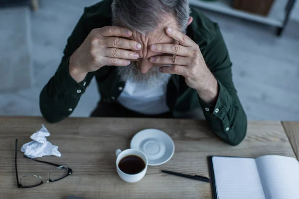 Blick aus der Vogelperspektive auf einen unglücklichen älteren Mann, der sich mit einer Tasse Kaffee und Schreibwaren auf einen Tisch lehnt — Stockfoto