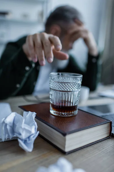 Blurred senior man reaching glass of whiskey on book near crumpled paper — Stock Photo