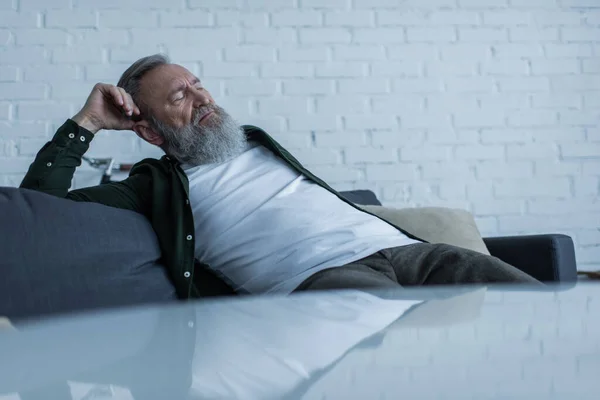 Molesto hombre mayor con la barba que sufre crisis mientras está sentado en el sofá en la sala de estar - foto de stock