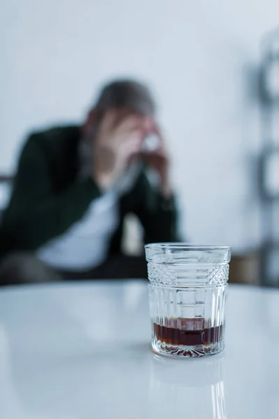 Glass of whiskey on white coffee table near blurred man suffering crisis — Stock Photo