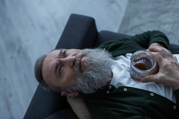 Вид зверху розчарованого старшого чоловіка з бородою, що лежить на дивані з келихом віскі під час кризи — стокове фото