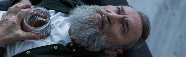 Vista dall'alto di uomo anziano deluso con la barba sdraiato sul divano con un bicchiere di whisky durante la crisi, banner — Foto stock