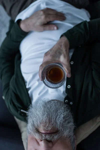 Обрезанный вид несчастного пожилого человека с бородой, лежащего на диване со стаканом виски во время кризиса — стоковое фото