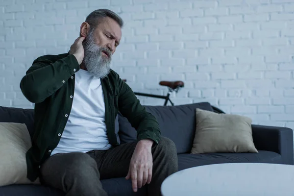 Unglücklicher älterer Mann mit Bart sitzt auf Couch und berührt Nacken, während er unter Schmerzen leidet — Stockfoto