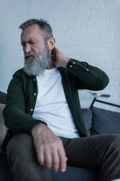 Upset senior man with beard and closed eyes sitting on couch and having neck pain — Stock Photo