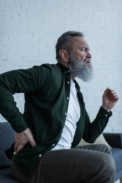 Bearded senior man with beard sitting on couch and touching back while suffering from pain — Stock Photo