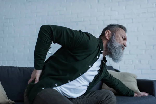 Homme âgé barbu avec barbe assis sur le canapé et touchant le dos tout en souffrant de douleur — Photo de stock