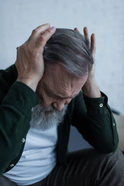 Estresado hombre mayor con barba tocando la cabeza mientras sufre de dolor de cabeza en casa - foto de stock