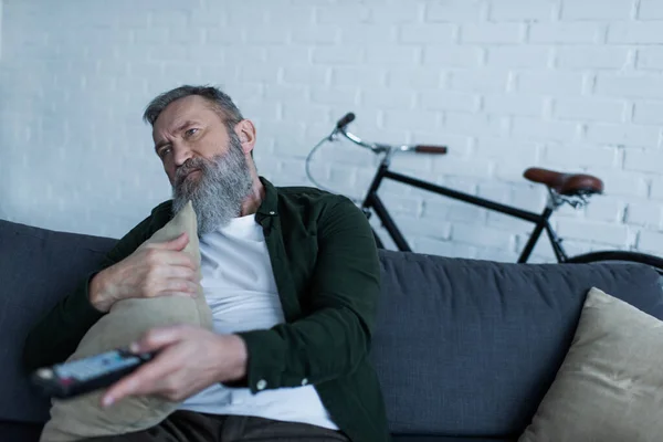 Uomo anziano depresso che tiene telecomando e cuscino mentre guarda la tv in soggiorno — Foto stock