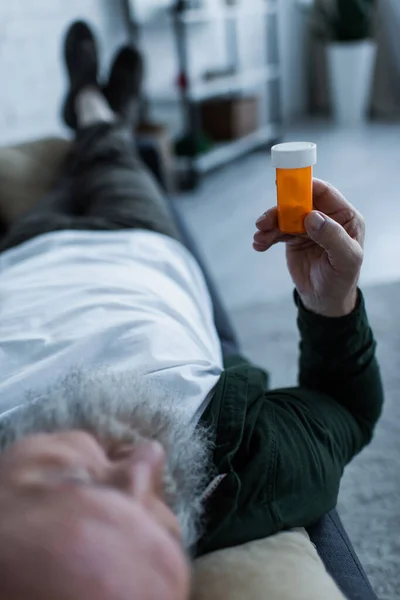 Розмитий старший чоловік тримає пляшку з антидепресантними ліками, лежачи на дивані у вітальні — стокове фото