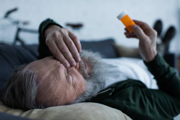 Hombre mayor deprimido sosteniendo la botella con medicamentos mientras está acostado en el sofá en la sala de estar - foto de stock