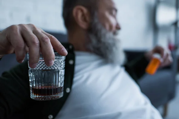 Blurred and unhappy senior man holding glass with whiskey and bottle with medication at home — Stock Photo