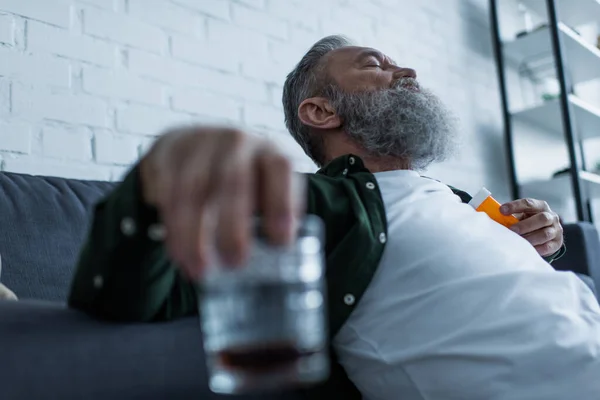 Бородатый мужчина с седыми волосами держит бутылку с лекарствами и стакан виски — стоковое фото